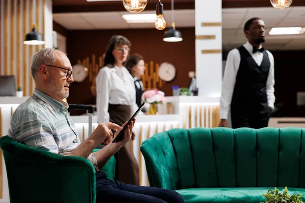 Old caucasian man using tablet to plan enjoyable holiday activities at fancy resort. In hotel lobby, elderly male guest using digital device while a staff member helps senior woman with her bags.