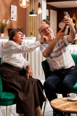 Yaşlı çift tatildeyken şık otel lobisinde görüntülü sohbet için mobil cihaz kullanıyor. Emekli kıdemli tatilciler dinlenme odasında, aile üyeleriyle akıllı telefonlardan sohbet ediyorlar..