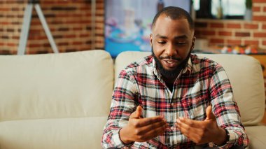 Modern şık bir apartman dairesinde dinlenirken arkadaşlarıyla çevrimiçi toplantı sırasında buluşacağı için heyecanlı gülümseyen neşeli insan. Video çağrısında arkadaşlarıyla sohbet eden bir adam