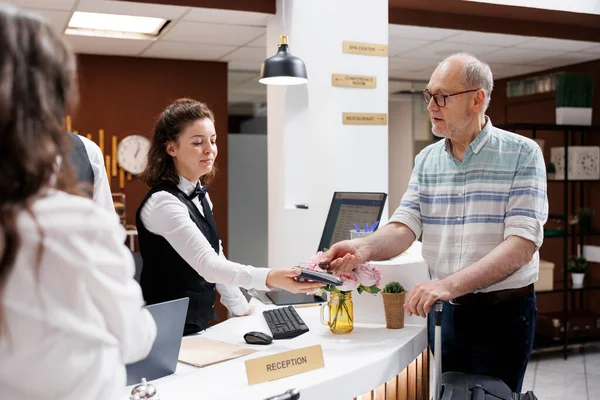 Retired senior man checks in at a hotel reception counter with the assistance of a concierge. Elderly male tourist pays with a credit card using an NFC terminal while holding his luggage.