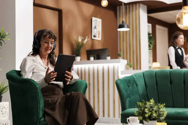 Elderly caucasian woman wearing headphones and sitting on sofa holding her tablet. Senior female guest having wireless headset waits in hotel lobby and uses digital gadget to browse the internet.