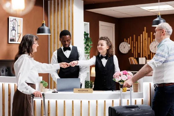 Friendly concierge assists elderly male and female tourists in hotel lobby. Retired senior woman provides necessary documents for check-in, while old man traveler waits patiently at registration desk.