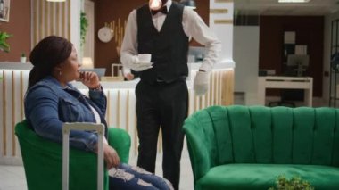 Porter bir fincan kahveyle servis yapıyor, lüks otelin dinlenme odasında oturuyor, bardan harika bir espresso servisi sunuyor. Misafir turist içecek alıyor.