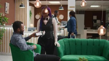 Gezgin otel personeline kahve fincanı için ödeme yapıyor, oda kaydını beklerken NFC mobil ödemesini pos 'ta yapıyor. Afrikalı Amerikalı müşteri içki ısmarlıyor, resepsiyonistle tartışıyor..