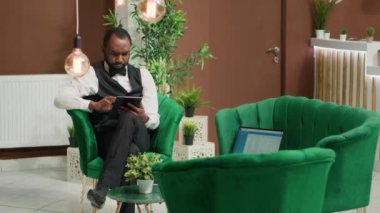 Lobide üniforma giyen Afro-Amerikan otel çalışanları online oda rezervasyonlarını yönetmek için dijital tablet kullanıyorlar. Belboy 'un elinde elektronik cihaz var. Yeni rezervasyonlar için ayrıntıları giriyor..