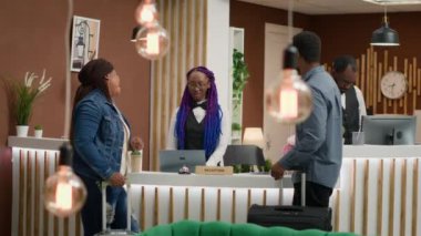 Otel kapıcısı beş yıldızlı otelde turistleri karşılıyor, kontrol için müşterilerle konuşuyor ve online oda rezervasyonunu gözden geçiriyor. Afro-Amerikalı insanlar bavullarıyla seyahat ediyor, resepsiyona geliyorlar..