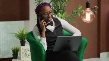 Afro-Amerikalı kadın resepsiyonist telefonla konuşuyor ve dinlenme odasında dizüstü bilgisayar rezervasyonu yaptırıyor. İnternet rezervasyonlarını yöneten otel çalışanı, telefon hizmeti.