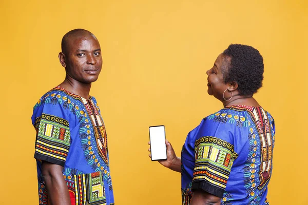 Smiling african american man looking at camera while woman holding smartphone with empty screen mockup for app. Black couple showing mobile phone with blank white touchscreen in vertical mode