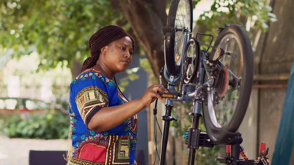 热情的黑人妇女仔细检查自行车轮子上的损坏以进行维修的肖像镜头 年轻的非洲裔美国妇女在院子里修理自己的自行车 检查踏板以修理 — 图库照片