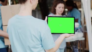 Beyaz bir kadın, elinde yeşil ekran krom anahtarıyla dijital tableti bir açık hava gıda bankasında yatay olarak tutuyor. Açık havada, hayır işi yapan bir kadın tarafından tutulan boş model şablonu gösteren akıllı bir cihaz..