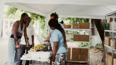 Kadın Afrikalı Amerikalı gönüllü, koltuk değnekli bir adama yemek ve ihtiyaçlarıyla yardım ediyor. Fakir, talihsiz ve evsiz insanlar yemek yardımı alıyorlar. Yan görünüm, el bilgisayarı.