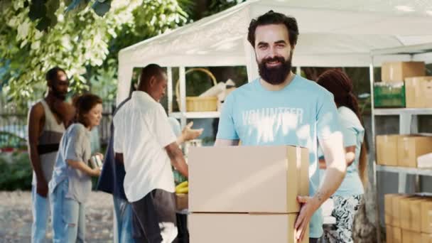近所の食糧運転でボランティアをしている寛大な男性は 飢えた人々やホームレスに食料を配布しています 寄付箱を持っている間 ワルカシアの慈善活動家はカメラを見ています トリポッドショット — ストック動画