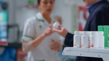 Eczane raflarındaki eczacılık ilaç şişelerinin yakın plan çekimleri. Eczacı bulanık arka planda müşteriye yardım ediyor. Eczanedeki tıbbi ürünler müşteriler tarafından satın alınmaya hazır.