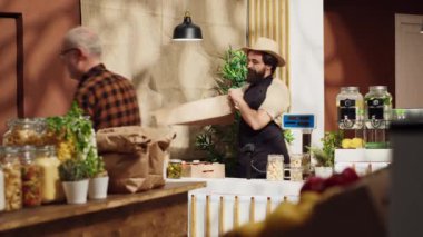 Sıfır atık süpermarket satıcısı. Müşteri alışverişini hazırlamak için kese kağıdı kullanıyor. Siparişi almak için teslimatçıyı bekliyor. Yerel mağaza satıcısı alışveriş torbalarına balkabağı ekliyor.
