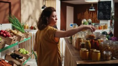 Sıfır atık süpermarketinden zımba alan kadın kasada parasını ödemeye devam ediyor. Yerel bakkaldaki müşteriler, yeniden kullanılabilir metal kavanozlarda büyük eşyalar satın alıyorlar.