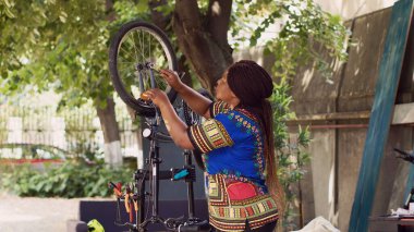 Motive olmuş siyah bir kadın ev bahçesinde bisiklet lastiğine ince ayar yapıyor. Profesyonel aletlerle. Genç Afrikalı Amerikalı bayan yaz tatili için bisiklet tekerleklerini yeniden diziyor ve emniyete alıyor..