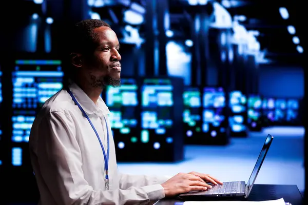 African american IT programmer in high tech facility using laptop to set up configuration management tools that enable automatic failover and load balancing, preventing server rigs system crash