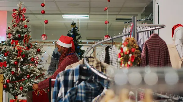African american employees wearing Santa hat ornating Christmas tree in clothing store before festive promotional event. Retail assitants ornating fashion shop during winter holiday season