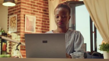 Sıcak ışıklandırılmış odada çalışan tele-işçinin portresi masa başında online rapor hazırlıyor, evdeki görevleri uzaktan çözüyor. Blogcu işi olan Afrikalı Amerikalı bir kadın. El kamerasıyla..
