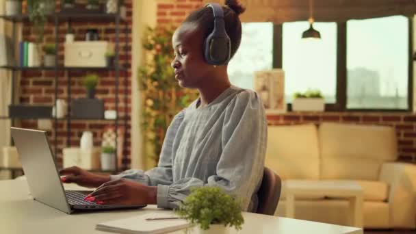Freelancerはデスクで音楽を楽しみながら オーディオヘッドセットで曲を聴きながらオンラインジョブタスクを楽しんでいる アフリカ系アメリカ人女性が日没でテレワークし ノートパッドでレポートを書く — ストック動画