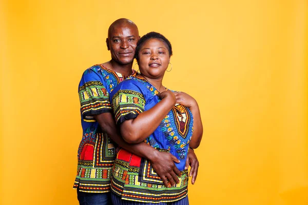 Mature black couple in love dressed in ethnic clothes embracing while posing for portrait together. Smiling husband hugging wife from back while standing and looking at camera