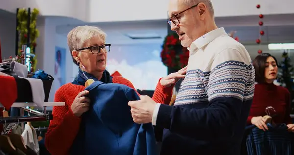 Senior couple browsing through clothes racks in festive adorn clothing store during Christmas shopping spree. Old clients planning to purchase formal attire, enjoying holidays promotional sales