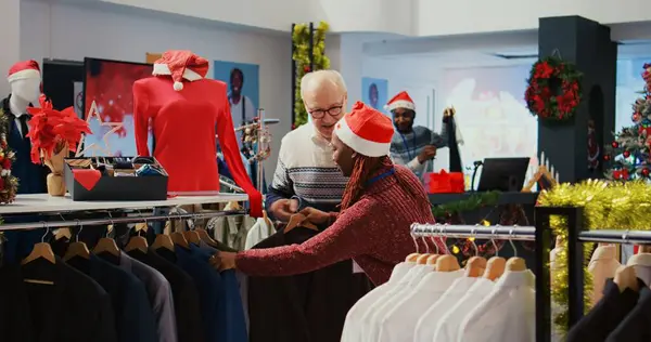 Elderly client being helped by retail assistant in festive decorated clothing store to determine if stylish blazer is the right fit. Employee assisting aged man in Christmas ornate fashion shop