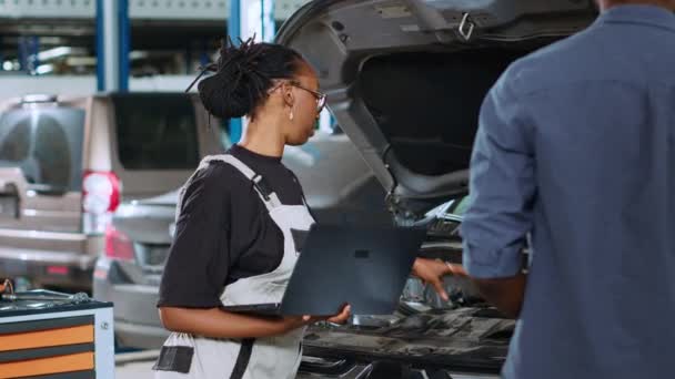 車の故障を修理した後 クライアントの修理請求書を計算するためにラップトップを使用して修理ショップのアフリカ系アメリカ人のメカニック 車両サービス後の最終コストを決定するデバイスを使用している女性 — ストック動画