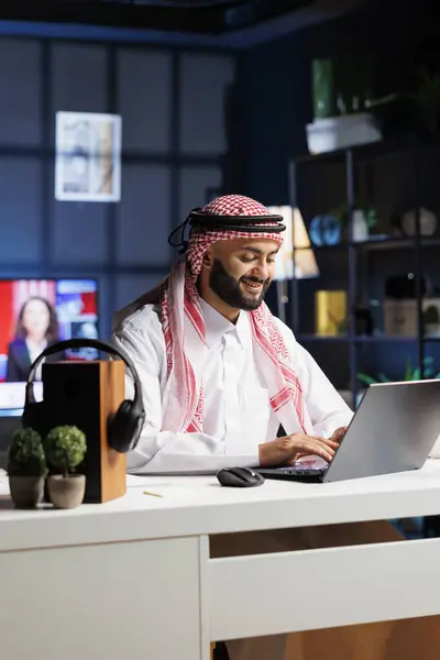 Arab businessman works diligently at his desk, typing on his digital laptop. Smiling Muslim guy using wireless technology for research and communication, showcasing his efficiency and professionalism.