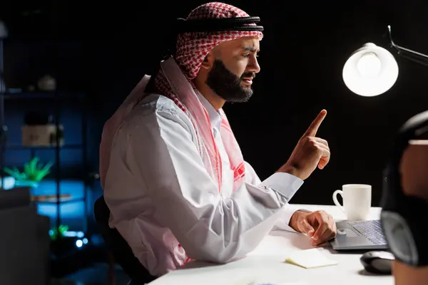 伊斯兰男子穿着传统衣服坐在办公桌前 笔记本电脑上有电话会议的侧视图照片 图为阿拉伯自由职业者在个人电脑上专心致志地交谈的情景 — 图库照片