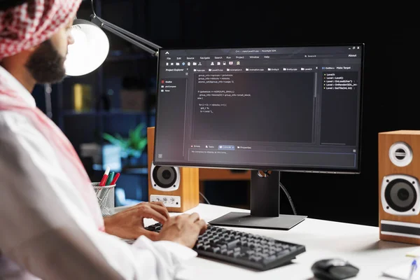 Muslim man sitting at desk and coding on computer, showcasing process of app development. Selective focus on pc monitor displaying running algorithms compiled by Arab guy in traditional attire.
