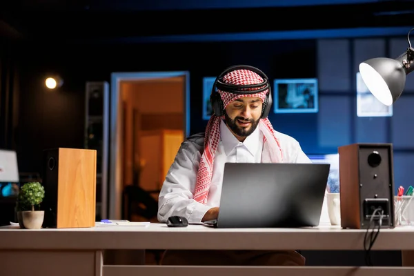 Confident Arab man typing on a laptop engaged in communication, video conferencing, and email. Muslim guy listens and watches with wireless headphones while researching and browsing the internet.