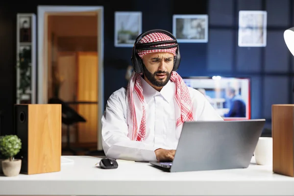 A traditional Middle Eastern entrepreneur diligently works in a modern office, utilizing wireless technology for communication and research. Young Arab man using his wireless headphones and laptop.