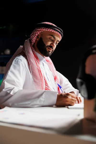 一名虔诚 专业的穆斯林男子在他的工作场所展示专业知识 一个穿着传统服装的中东人在笔记本上写下的详细侧面照片 — 图库照片
