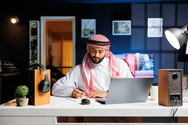 Arab man works efficiently browsing the internet, takes notes, and communicates digitally in his modern office. Young Muslim guy doing research and writing on his notebook.