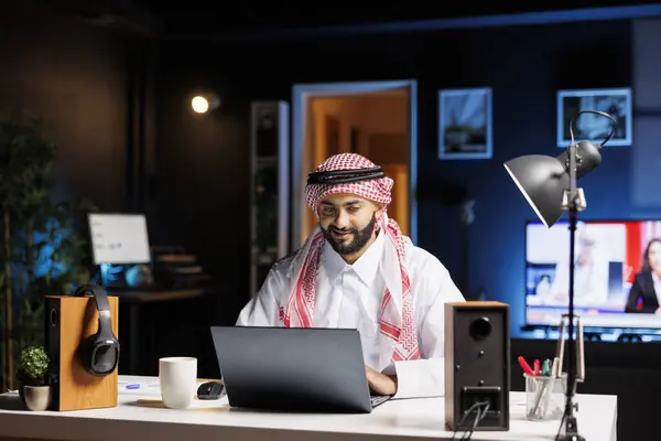 Focused man in Arabic attire working diligently at his modern office desk, typing on his laptop. Muslim guy surfing the net and checking his e-mail on his digital personal computer.