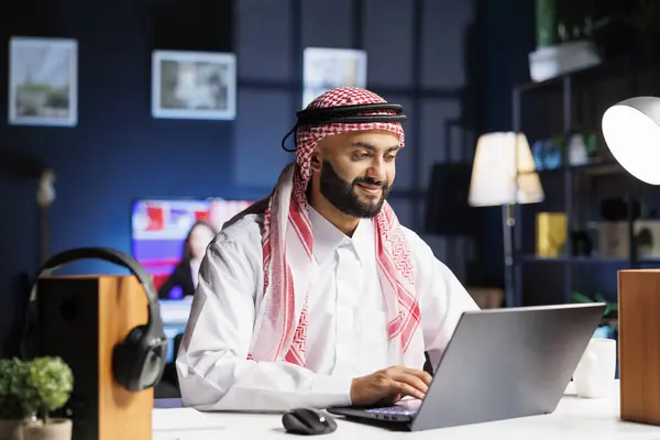 Young Arab man works diligently at his desk, browsing the internet on his digital laptop. Engrossed in the online webpage, he takes notes and researches, embodying professionalism and efficiency.