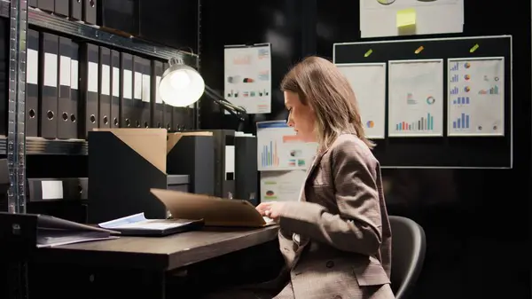 Caucasian female inspector examines confidential case files and records. Young private detective walks into office space filled with clues and evidence, positioning laptop on table and starts working.