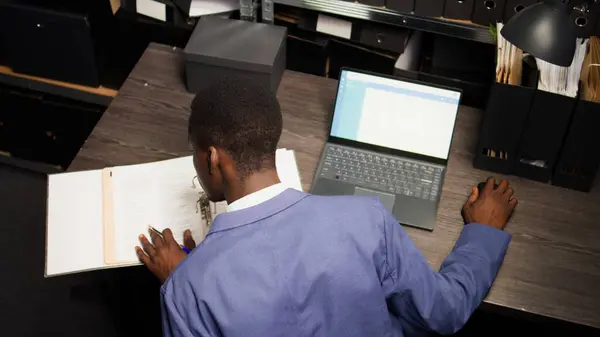 Male inspector observed diligently analyzing files and evidence acquired during research in archive room. Law officer uses laptop to cross-reference records and do a background check. Aerial shot.