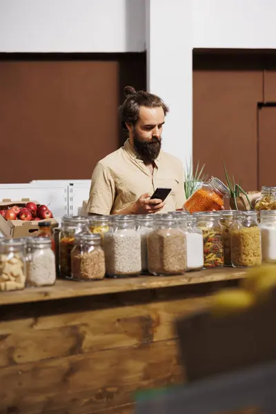 Man in zero waste store analyzing pantry products, using smartphone to make sure they are freshly harvested. Vegan customer thoroughly checking local supermarket food items are preservatives free