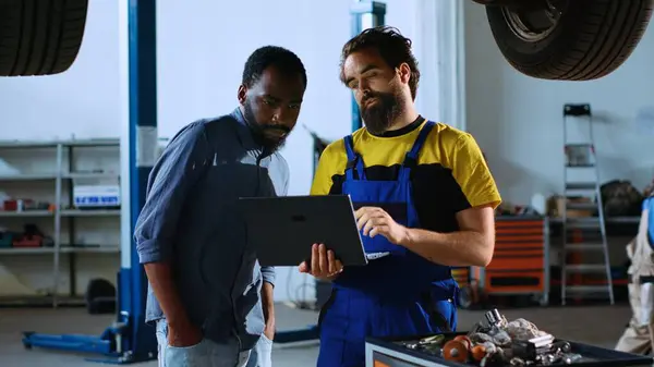 Licensed repairman in garage workspace standing with customer underneath suspended car, looking together for replacement parts. Worker helps man fix vehicle, using laptop to find components