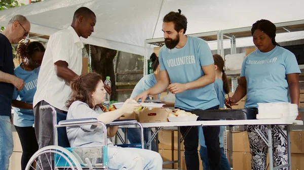 Helpful guy volunteer offers free lunch to the poor disabled woman in need. Multiracial charity workers at an outdoor food bank provide the handicapped and less fortunate with hunger relief.