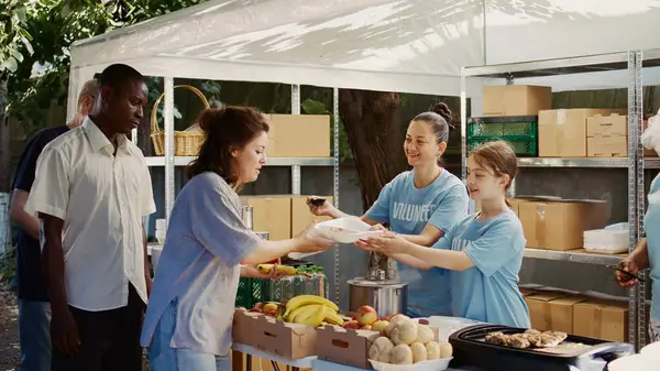 Caucasian girl packs free food to be distributed to the underprivileged with assistance from other charity workers. Female volunteers serving hot meals to the homeless and hungry at outdoor food bank.