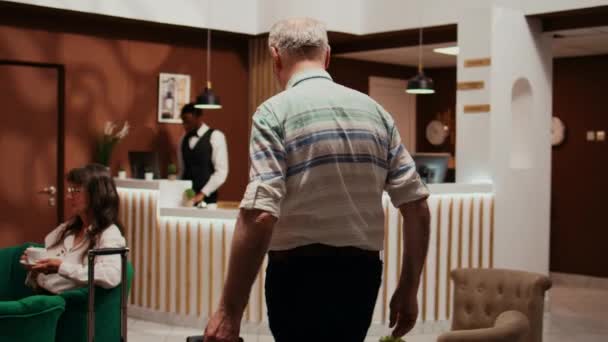 ホテルのフロントにトロリーバッグを持って到着する高齢者は チェックインの手伝いを求めます すべての包括的な休日のリトリート 観光産業で老人にとって快適な滞在を確保するレセプション — ストック動画