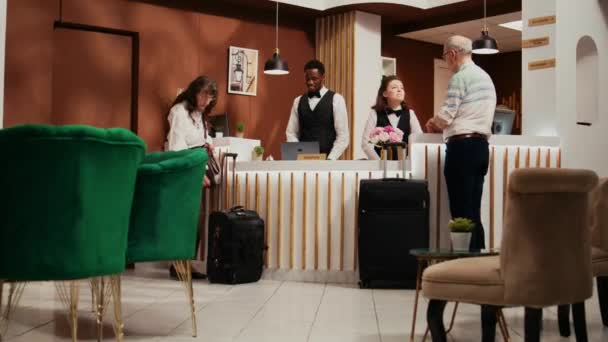 フロントスタッフが5つ星ホテルの客室予約のためのパスポートをチェックし 受付ロビーでId文書を見ています シニアカップルを支援する多様な従業員 — ストック動画