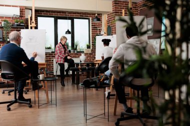 Çeşitli yaşlarda, ressamlarla birlikte resim yapmayı öğrenen, grup sanatları dersine katılan, sınıfta kalemle resim çizen bir grup farklı insan.