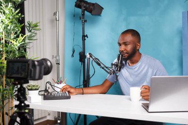 Vlogger internet kanalı için video kaydederken ses kontrol cihazı kullanıyor ve kahve içiyor. Afrikalı Amerikalı adam ses karıştırıcı ile blog için dijital içerik yaratıyor