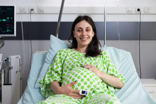 Smiling pregnant woman lying in bed holding hands on belly in hospital ward. Cheerful patient with belly pregnancy looking at camera and waiting on doctor to do examination during checkup visit
