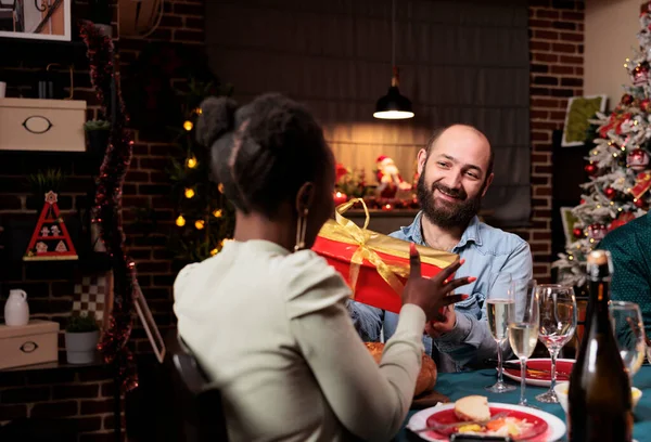 Diverse friends offering gifts to each other for christmas holiday, spreading positivity at festive dinner. Happy man giving present box to african american woman during winter celebration.