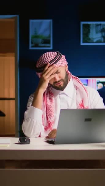 垂直ビデオ 疲れたアラブのビジネスマンは 自宅のオフィスで働く片頭痛に苦しんでいます リビングルームで疲れ果てた中東の従業員は 過労から頭痛を得る電子メールに答えます — ストック動画